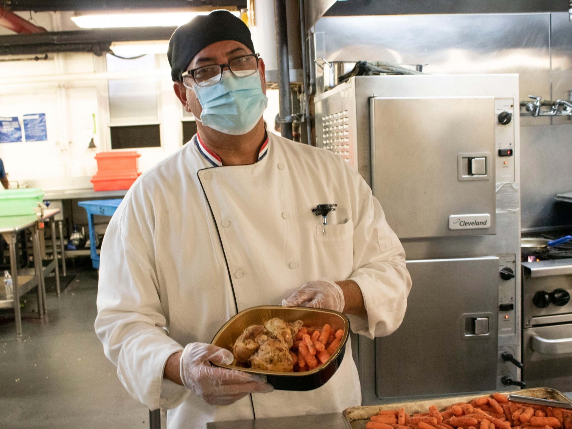 Chef Mike standing the kitchen holding a to-go box of roasted chicken and carrots.