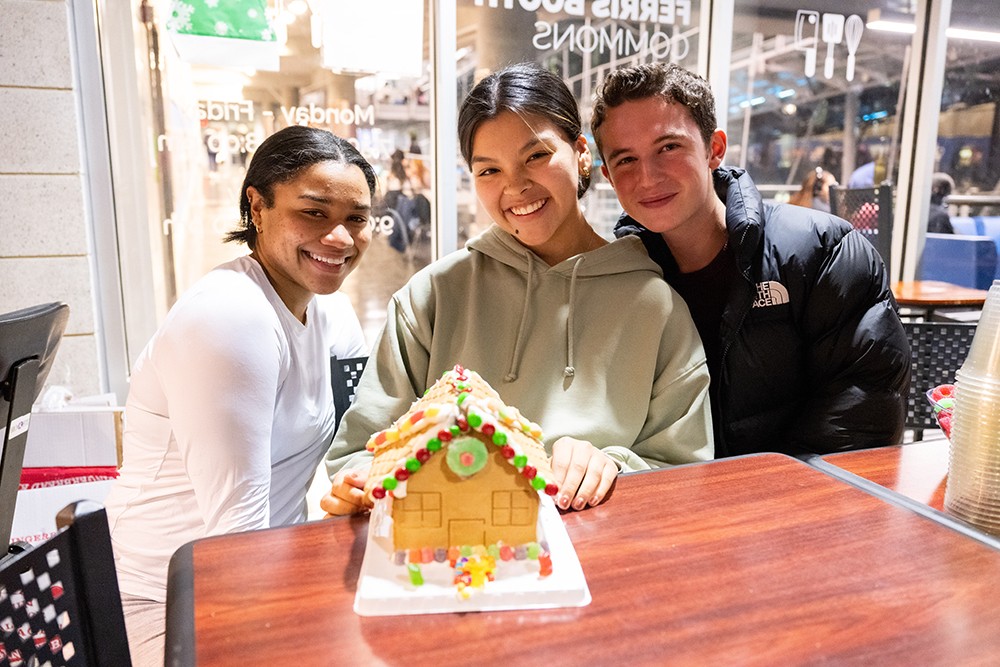 Three students sit at a table after decorating a gingerbread house