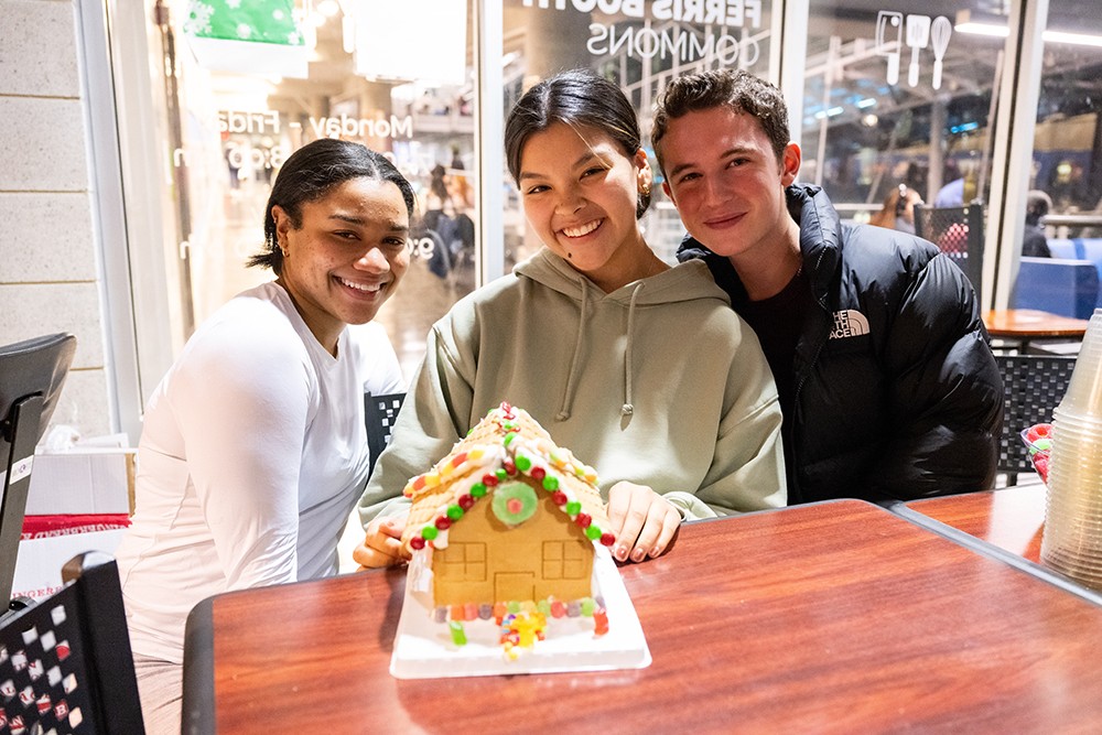 Are you architecture students? Because that gingerbread house is a masterpiece. Three students sit at a table after decorating a gingerbread house