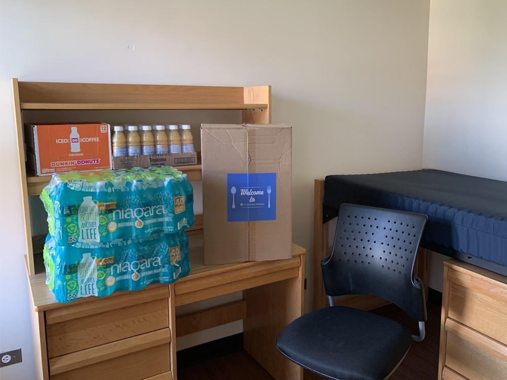 Welcome boxes delivered to all student’s room in advance of Check-In, along with a supply of water, juice, and coffee. (Photo: Campus Services) A dorm room that has a Welcome Box along with a supply of water, juice, and coffee sitting on a desk.