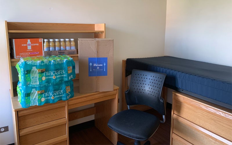Cases of bottled water, orange juice, and iced coffee sit on a desk in a dorm room
