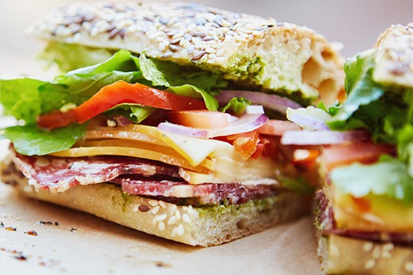 Close up of sandwich on a seed bun with lettuce, tomato, cheese, pesto sauce, and salami