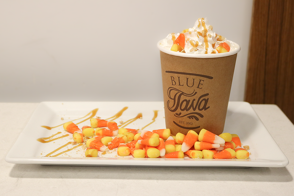 A Blue Java beverage cup topped with whipped cream, caramel drizze and candy corn sits on a plate. The plate is dressed with candy corn and a caramel drizzle.