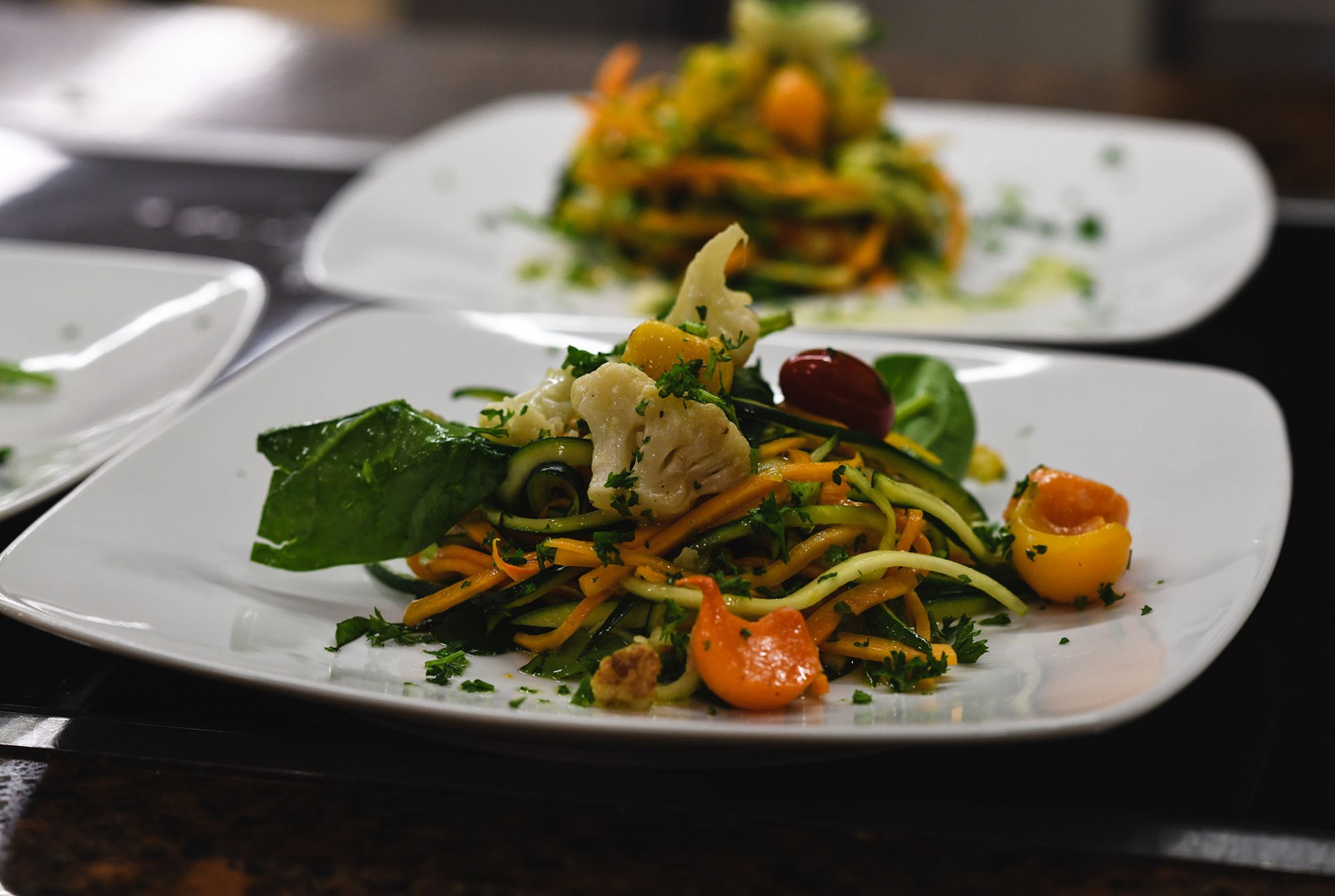 Veggie salad with tomatoes, lettuce, cauliflower, carrots, and garnish plated on a plate. Out of focus veggie salad is behind the dish in focus.